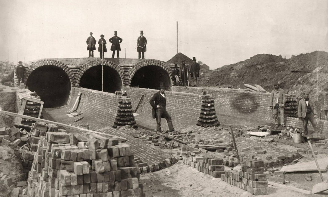 Historic black-and-white photo showing workers building brick tunnels, with men in suits and hats standing on top. Piles of bricks and construction materials are visible around the site.