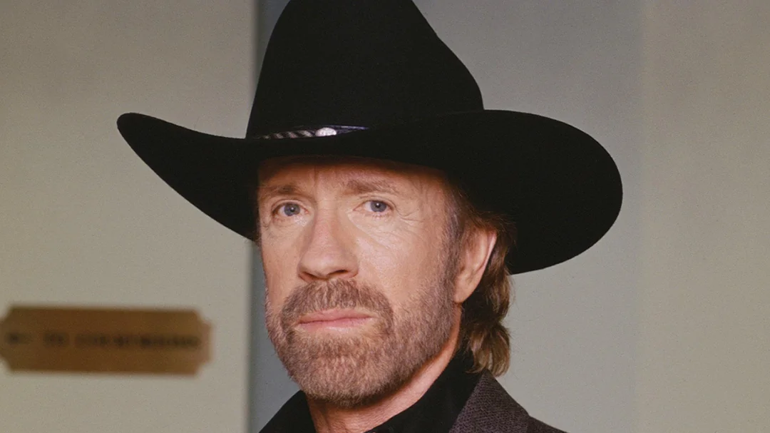 A man with a beard and mustache wearing a black cowboy hat and dark jacket stands indoors in front of a blurred background.