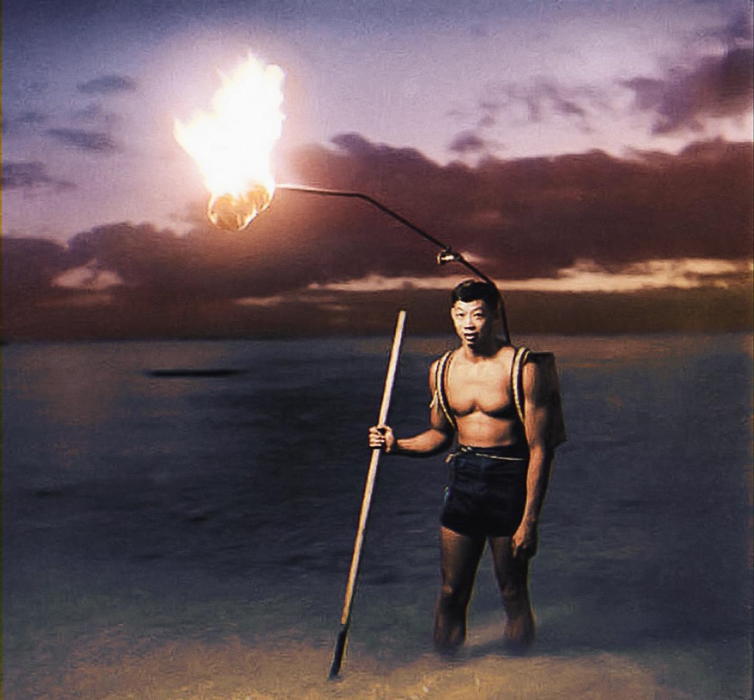A man stands in shallow water at dusk, wearing shorts and a harness with a tall pole attached to his back, holding a flaming torch above his head. The sky is dramatic with dark clouds and a purple-orange sunset.