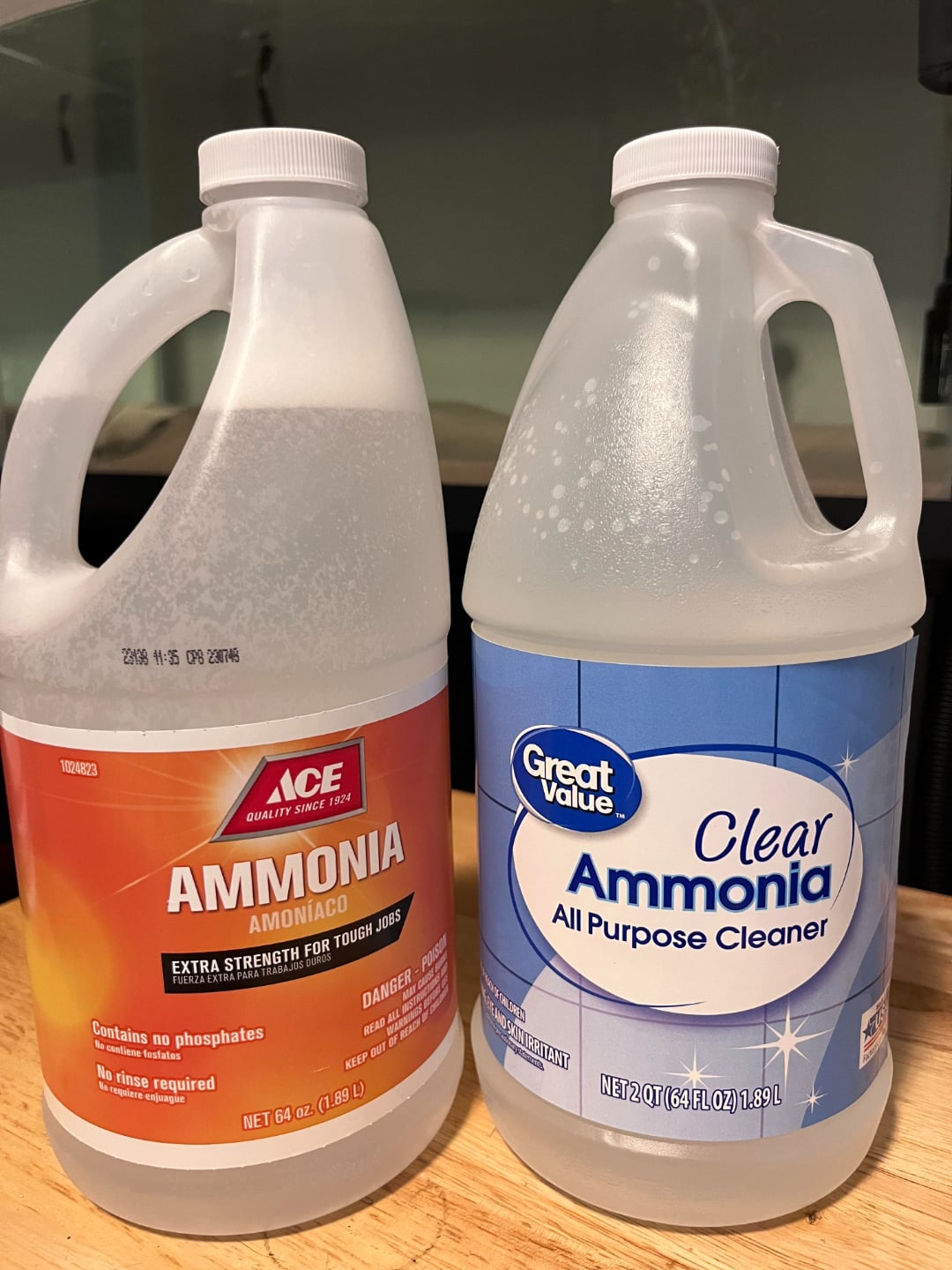 Two one-gallon bottles of ammonia are on a wooden surface. The left bottle is Ace brand with an orange label, and the right bottle is Great Value brand with a blue and white label. Both are labeled as cleaners.