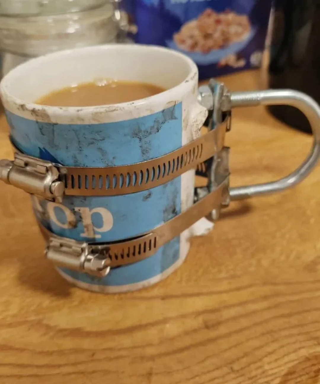 A chipped blue and white mug filled with coffee has a makeshift handle made from two metal hose clamps and a metal bracket, sitting on a wooden surface.