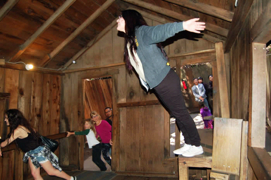 A woman leans forward at a dramatic angle in a tilted wooden room, arms outstretched as if balancing. Other people inside appear to be struggling to stand upright due to the slanted floor and walls.