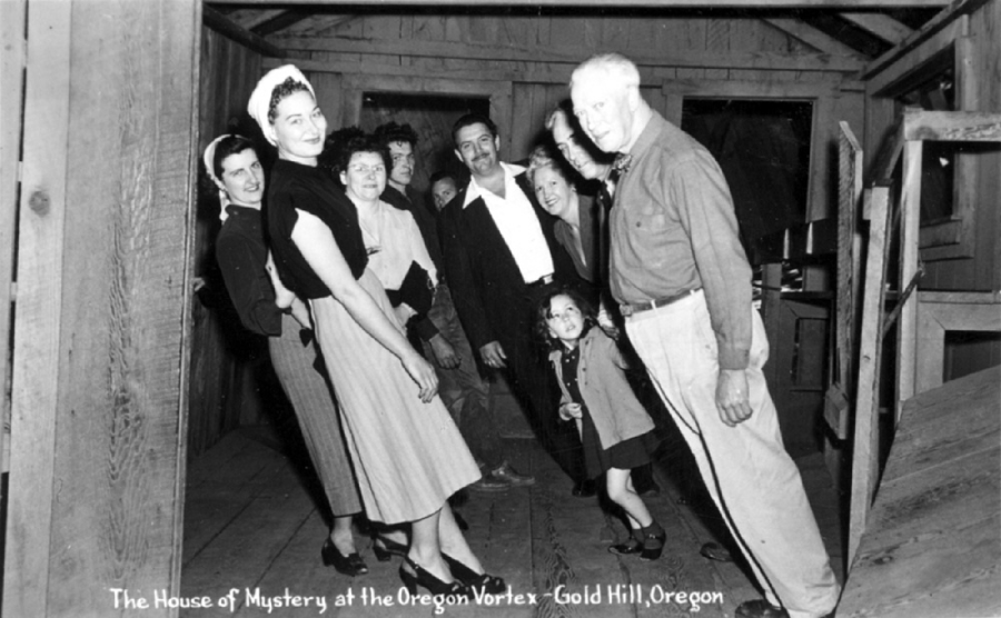A group of people lean at odd angles inside a tilted wooden room at The House of Mystery, Oregon Vortex, Gold Hill, Oregon. The walls and floor create an optical illusion of imbalance.