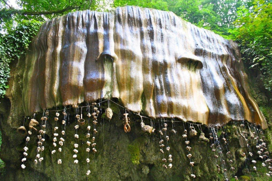 A large, mossy rock formation with a smooth, draped appearance, resembling a waterfall. Various ceramic faces, shells, and objects hang from strings underneath, surrounded by green foliage.
