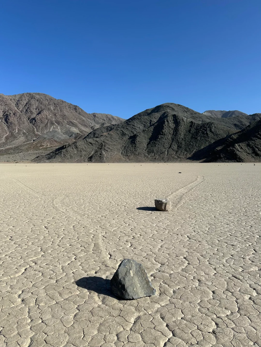 Large rocks sit on a cracked, dry desert floor, each leaving a long trail behind them, with dark mountains and a clear blue sky in the background.