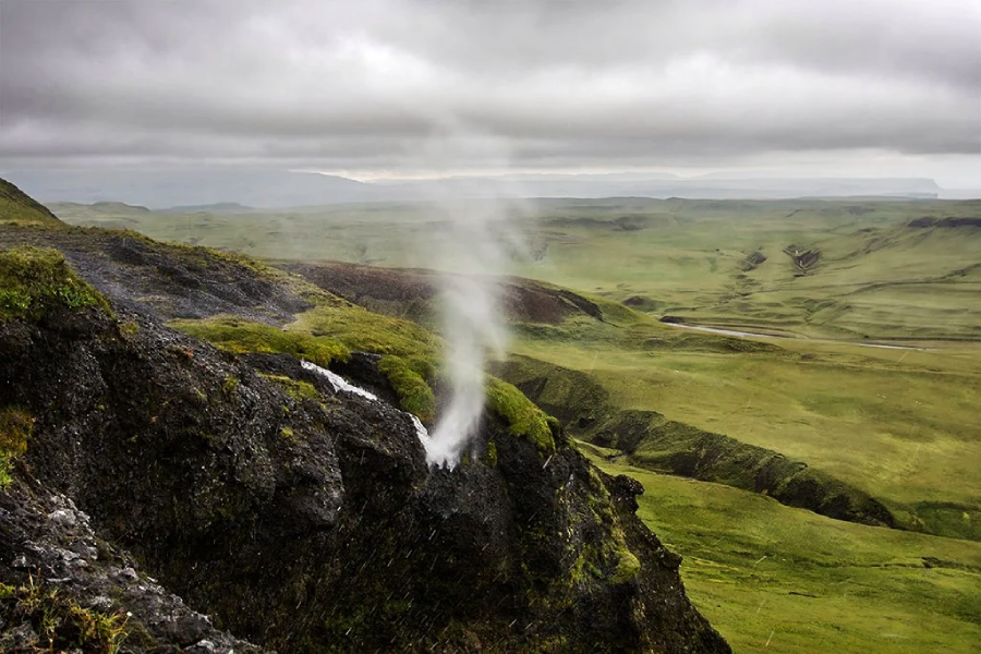 Steam rises from a rocky, moss-covered hillside overlooking a wide, green valley under a cloudy, overcast sky. The scene appears remote and natural, with rolling hills in the distance.
