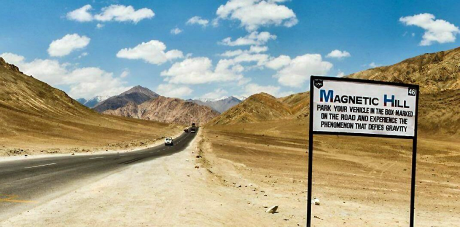 A road winds through barren, rocky mountains under a blue sky with scattered clouds. A sign on the roadside reads “Magnetic Hill: Park your vehicle in the box marked on the road and experience the phenomenon that defies gravity.”
