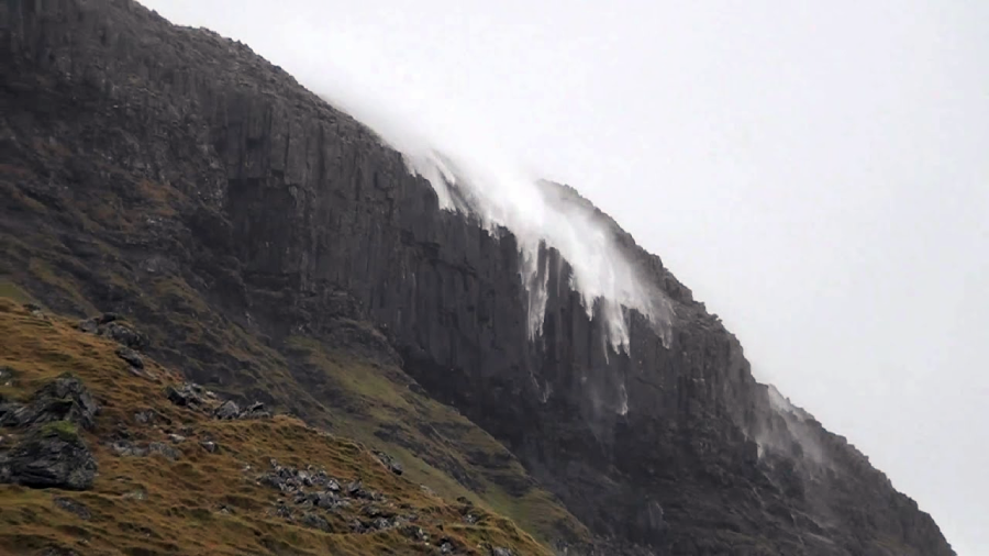 Strong winds blow a waterfall upward against a steep, rocky mountainside with patches of green moss or grass, creating a dramatic, misty effect in overcast weather.
