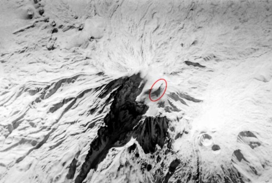 Aerial view of a snow-covered volcanic landscape with dark ash trails radiating from the crater; a red circle highlights a specific area near the volcano’s summit.