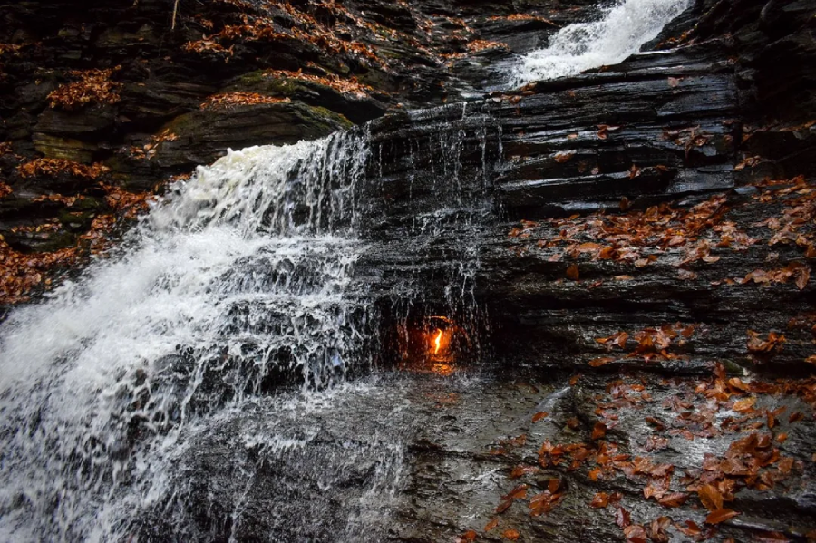 A small waterfall flows over dark rocks covered with autumn leaves. Behind the falling water, a small orange flame is visible, creating a unique natural scene.