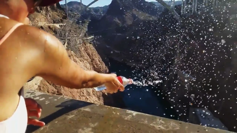 A person in a tank top squirts water from a bottle, creating a spray of droplets in the air, with a rocky canyon and a bridge visible in the background.