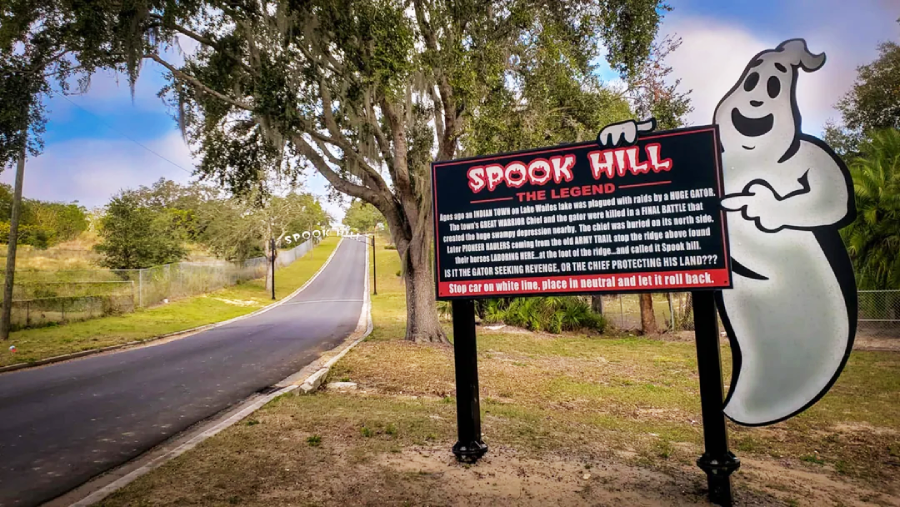 A road stretches uphill with a large sign in the foreground reading "Spook Hill: The Legend," next to a smiling cartoon ghost and shaded by large trees.