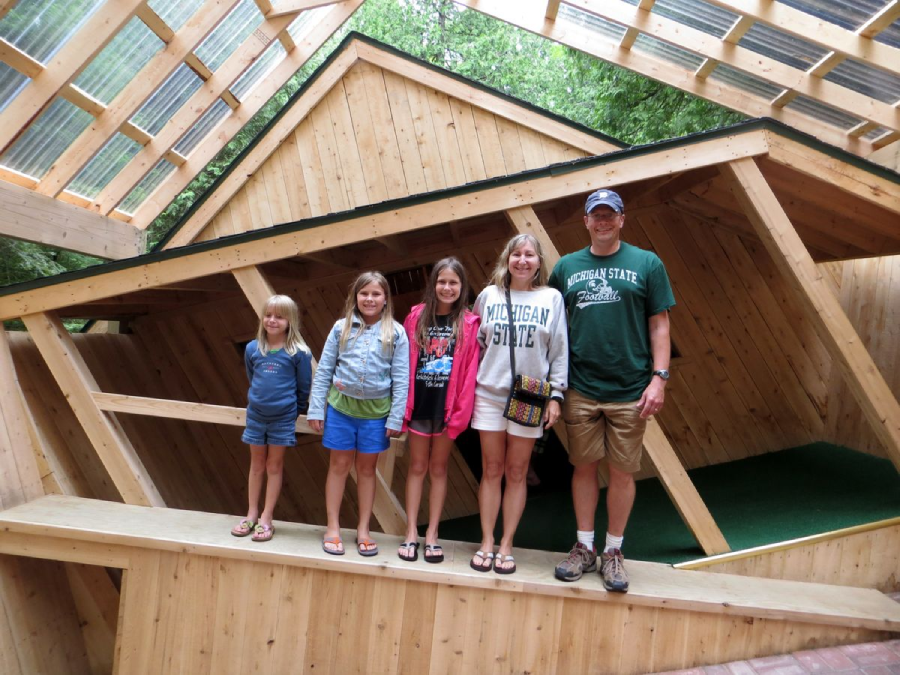 Five people stand on a slanted wooden floor inside a tilted wooden house, under a transparent roof. The group includes three children and two adults, all smiling at the camera. Trees are visible in the background.