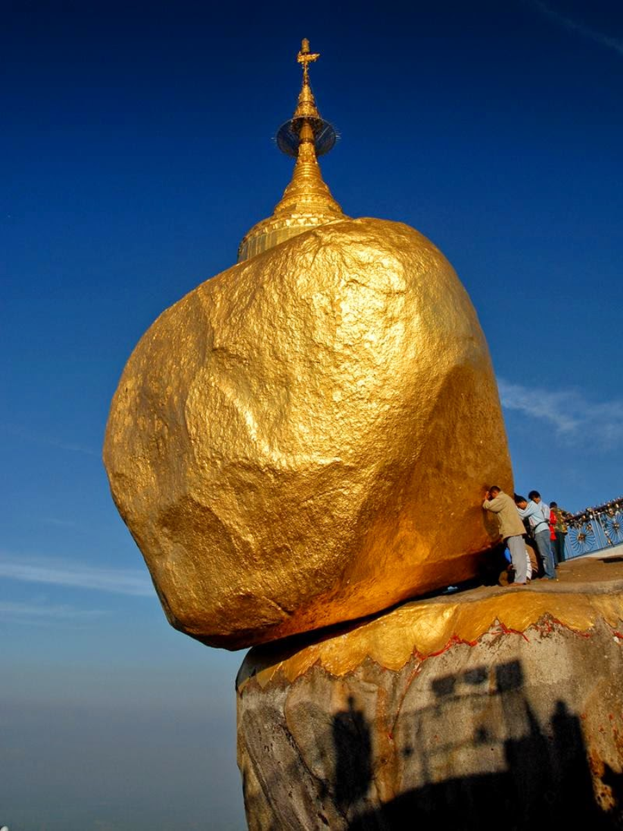 A large golden rock, known as the Golden Rock Pagoda, balances on the edge of a cliff under a clear blue sky, with people standing nearby for scale.