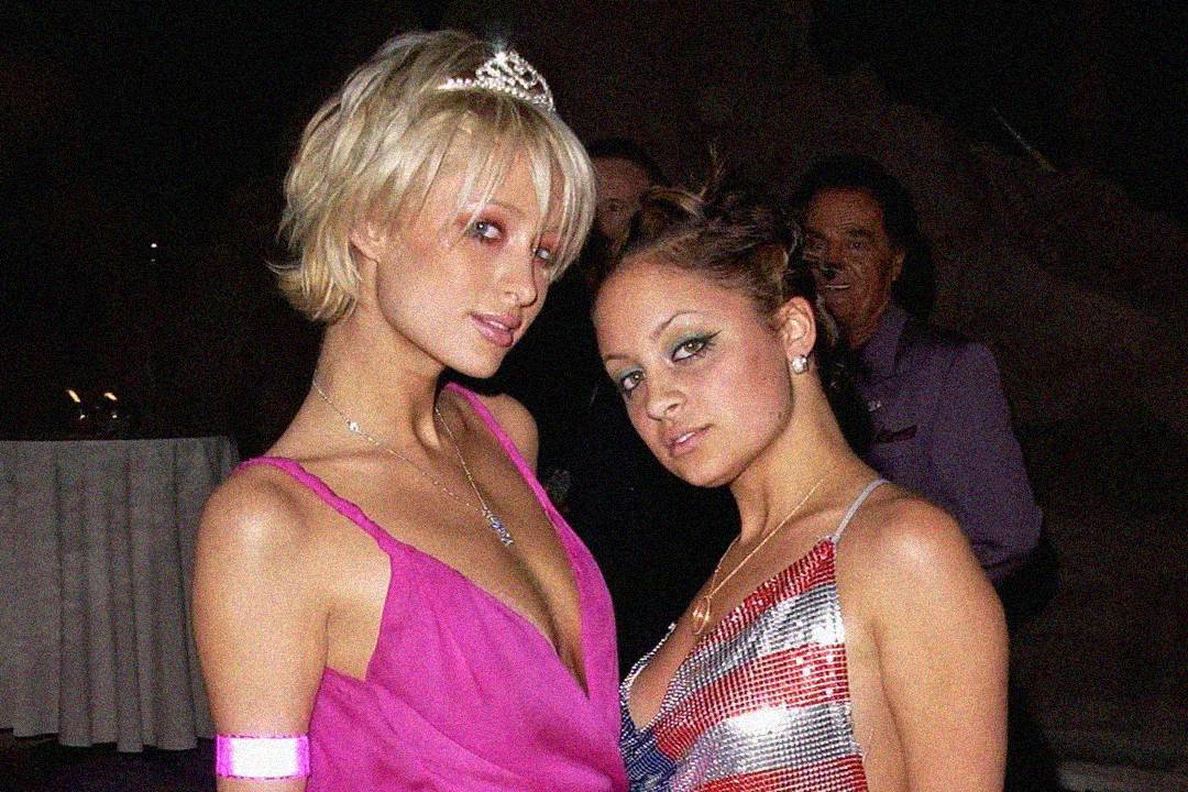 Two women pose closely together at a nighttime event. One wears a pink dress and a tiara, the other a silver dress with bold eye makeup. The background is dimly lit with a man and a table visible.