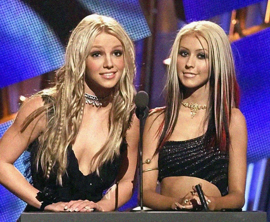 Two young women with long blonde hair stand at a microphone onstage. Both wear black outfits with sparkly details and chokers, appearing at an awards show with a colorful, lit backdrop behind them.