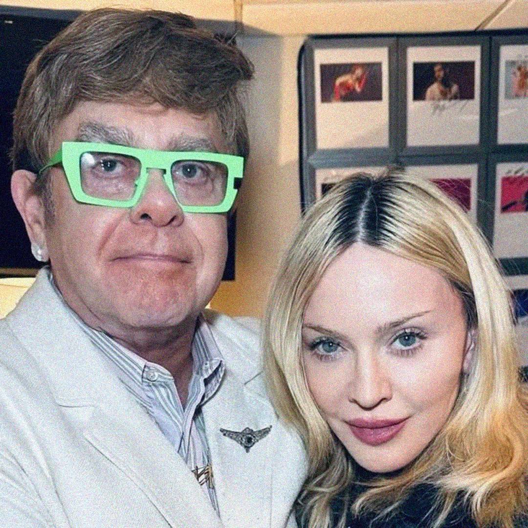 A man with light brown hair and bright green glasses stands next to a blonde woman; both are facing the camera and smiling slightly. There are photos displayed on the wall in the background.