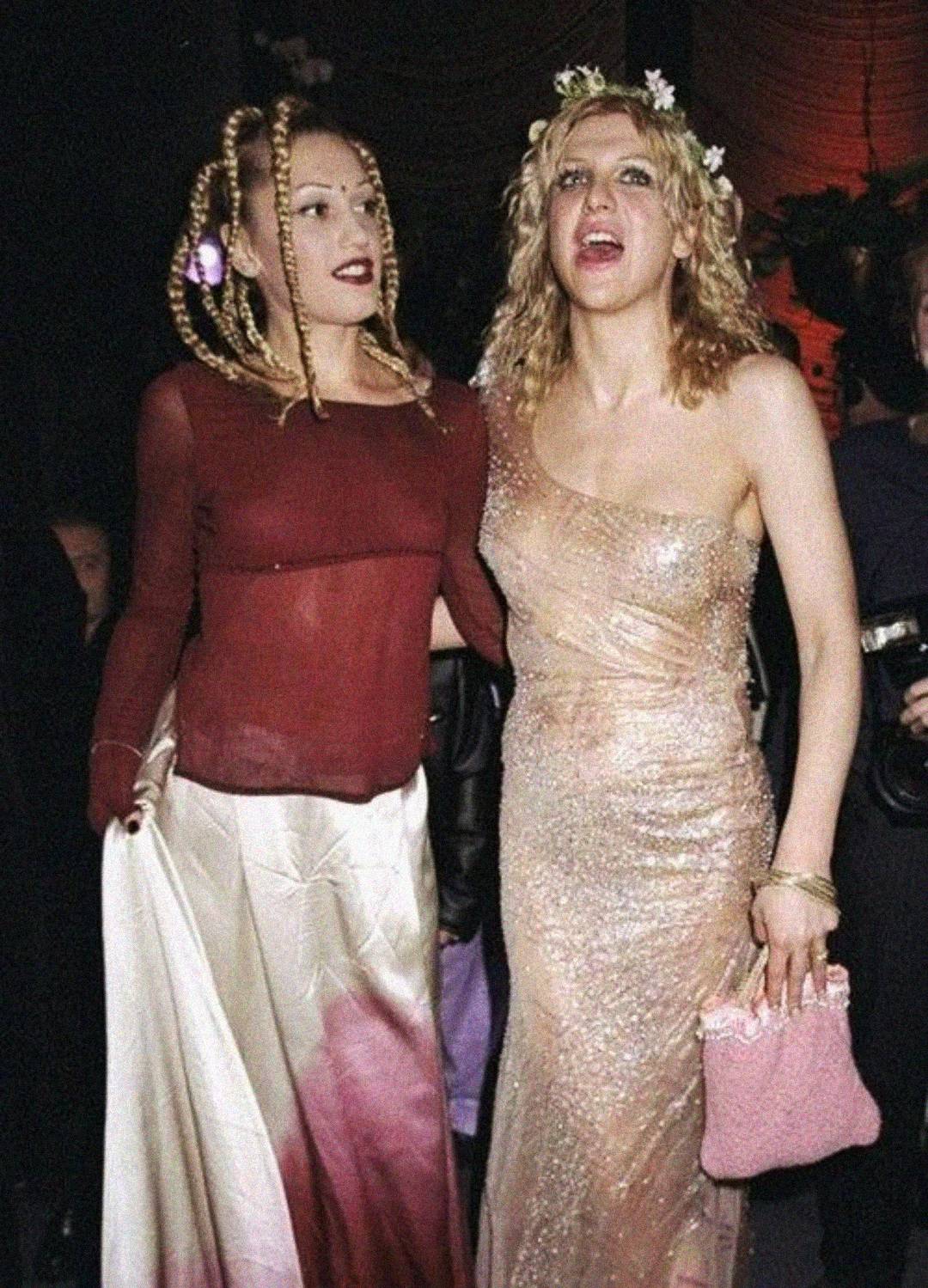 Two women pose together at an event. One wears a sheer red top with a long white-pink skirt and has braided hair. The other wears a sparkly, one-shoulder beige gown with a pink purse and floral crown in her wavy hair.