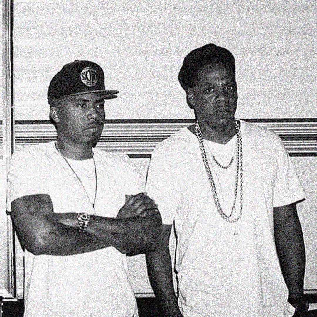 Two men wearing white t-shirts and chains stand side by side. One has arms crossed and wears a cap, while the other wears a beret-style hat. The photo is black and white, and both have serious expressions.