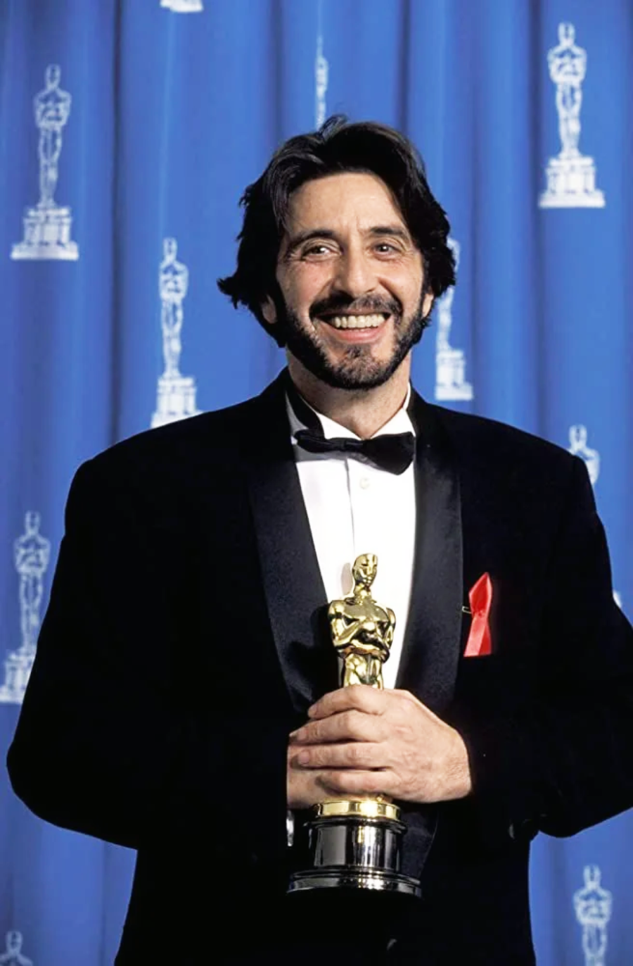 A man in a tuxedo smiles while holding an Oscar statuette. He stands in front of a blue curtain with Oscar logos and wears a red awareness ribbon on his lapel.