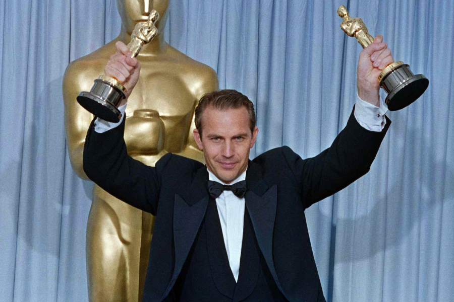 A man in a tuxedo holds up two Oscar statuettes triumphantly, standing in front of a large gold Oscar statue and blue curtains.