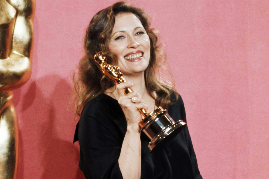 A smiling woman with wavy hair holds up an Oscar trophy in front of a pink backdrop. She is wearing a black outfit and standing next to a large golden Oscar statue.