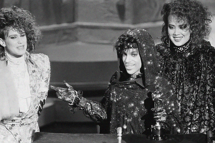Three people with curly hair stand together on stage in glamorous outfits. The person in the center, wearing a sparkly hooded cape, holds an Oscar statuette and gestures with their hand, while the others smile.