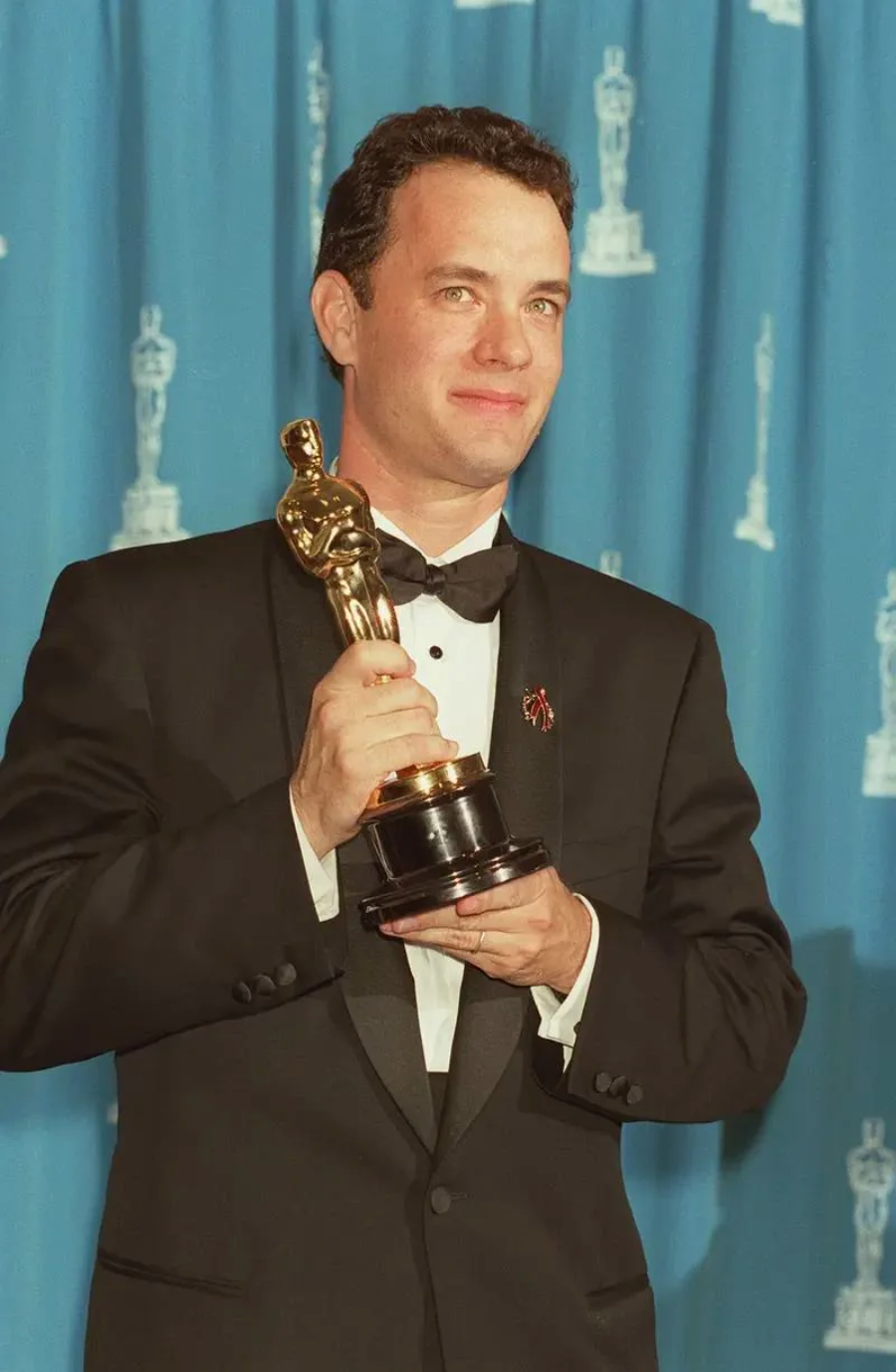 A man in a black tuxedo holds an Oscar trophy and smiles while standing in front of a blue curtain decorated with Oscar statuette patterns.