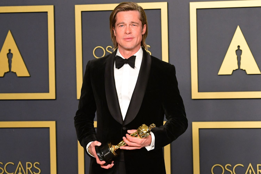 A man in a black tuxedo holds an Oscar trophy and poses in front of a backdrop with Oscar logos at an awards event.
