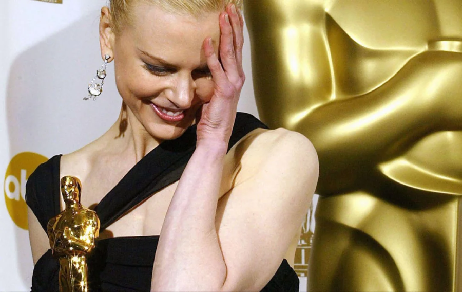 A woman in a black dress smiles shyly and touches her face while holding an Oscar statuette, standing in front of a large gold Oscar statue at an awards event.