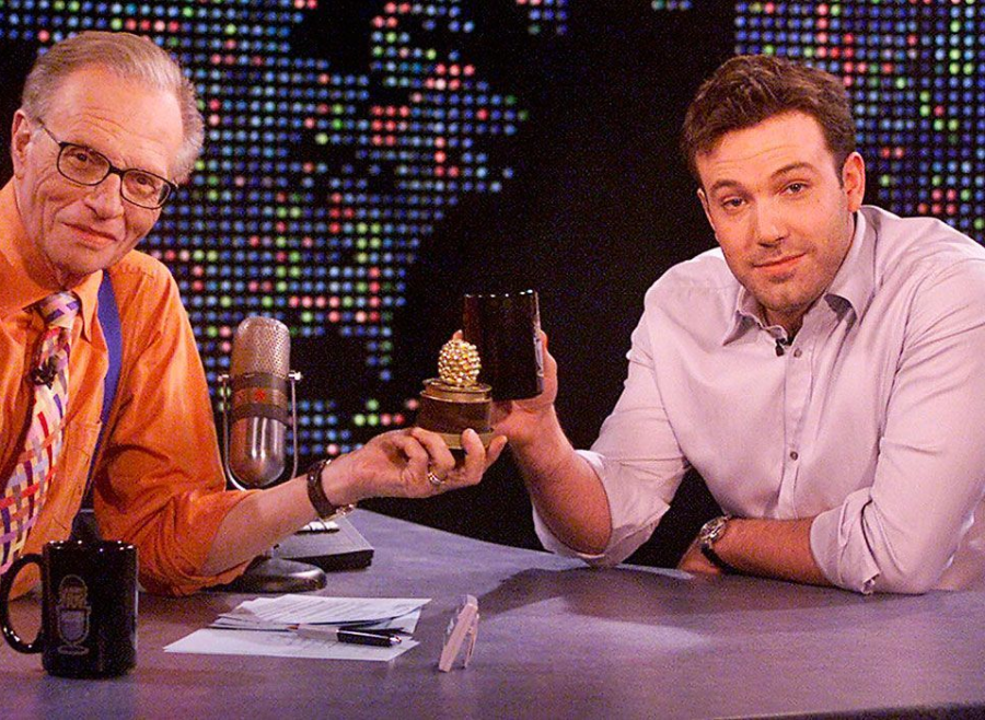 Two men sit at a desk holding a small golden trophy and a black mug, smiling at the camera. Colorful lights form a dotted world map in the background. One man wears glasses and an orange shirt; the other wears a light shirt.
