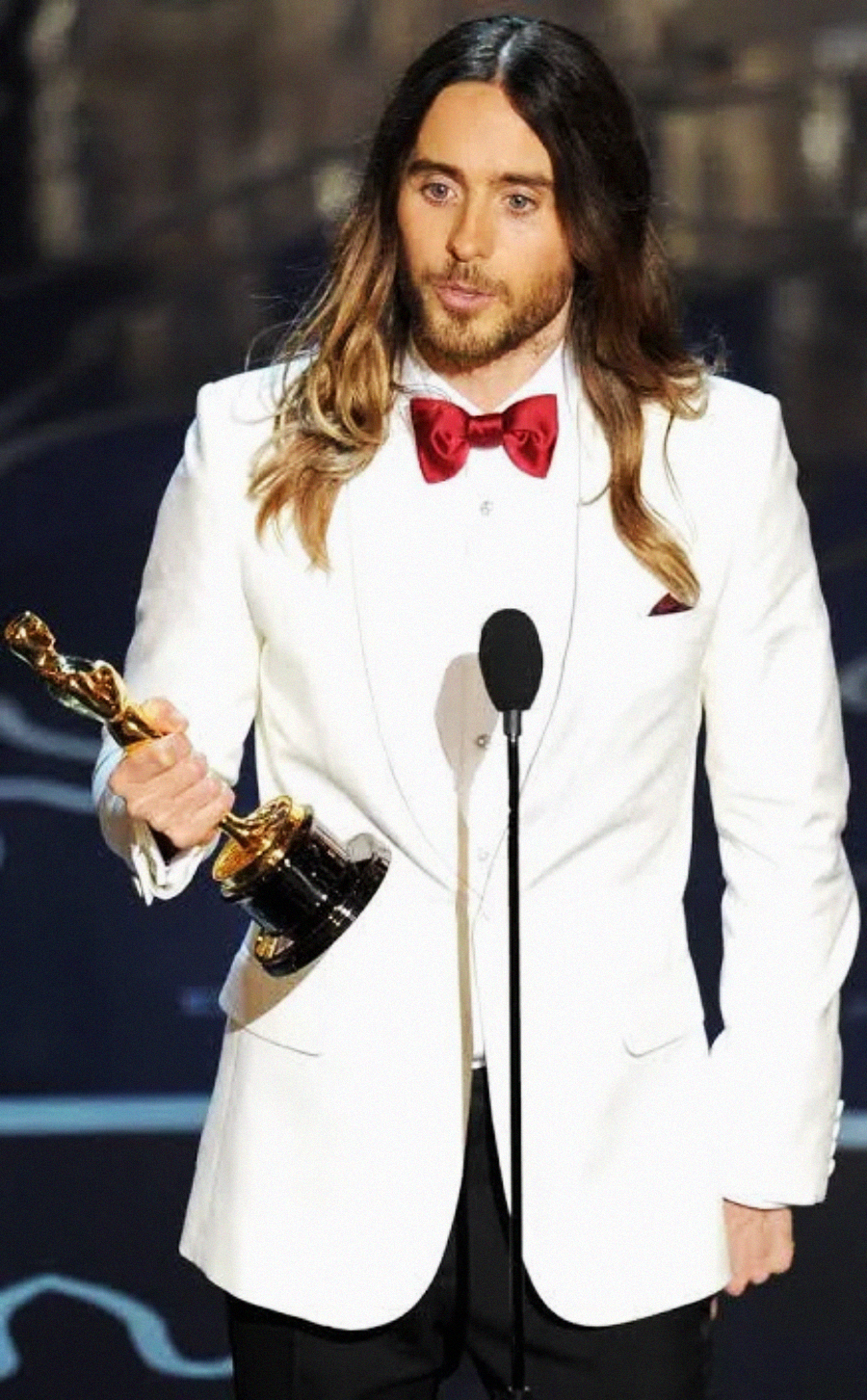 A man with long brown hair and a beard, wearing a white suit jacket, red bow tie, and black pants, stands at a microphone holding an Oscar trophy.