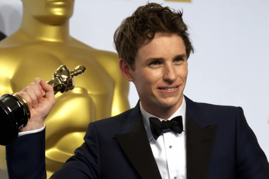A man in a navy tuxedo and black bow tie smiles while holding up an Oscar trophy, standing in front of a large gold Oscar statue.