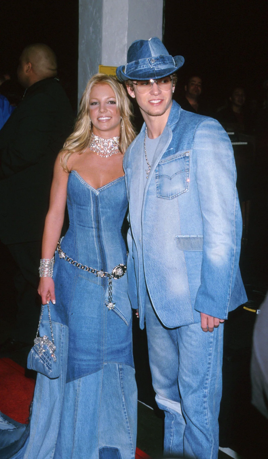 A woman and a man pose together on a red carpet, both wearing matching blue denim outfits, including a dress, suit, and hat. The woman carries a denim purse and wears silver jewelry.