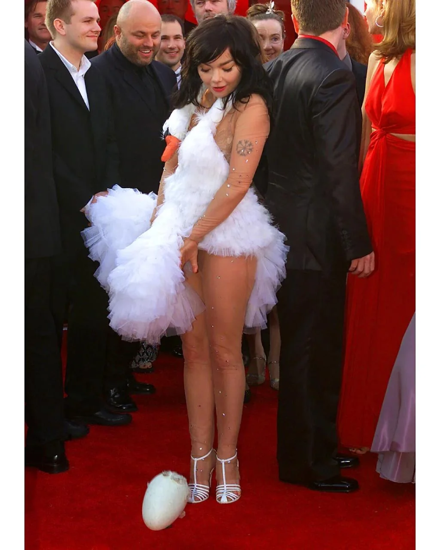 A woman in a white swan-inspired dress stands on a red carpet, holding part of her dress and looking down as an egg-shaped object lies at her feet. People in formal attire stand around her.