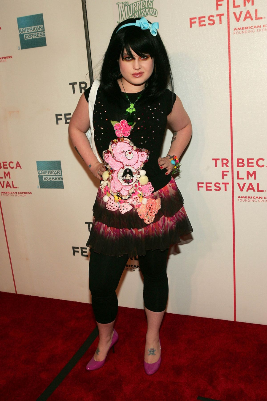 A person with black hair, a blue bow, and colorful clothes featuring a pink cartoon bear poses on the red carpet at the Tribeca Film Festival. The background has event logos and sponsor signs.