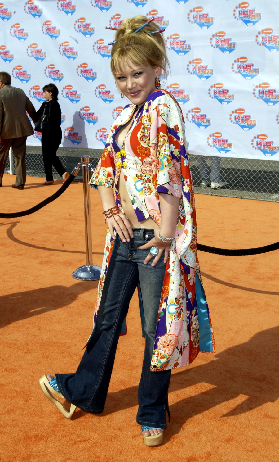 A young woman poses on an orange carpet, wearing flared jeans, a colorful long kimono-style jacket, platform sandals, and styled blonde hair. The background features a Nickelodeon Kids’ Choice Awards step-and-repeat banner.