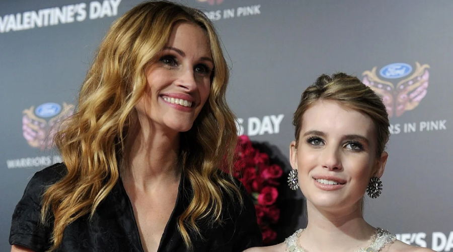 Two women stand together smiling at an event. The woman on the left has long, wavy blonde hair and wears a black outfit. The woman on the right has light brown hair in an updo and wears a light-colored dress with statement earrings.