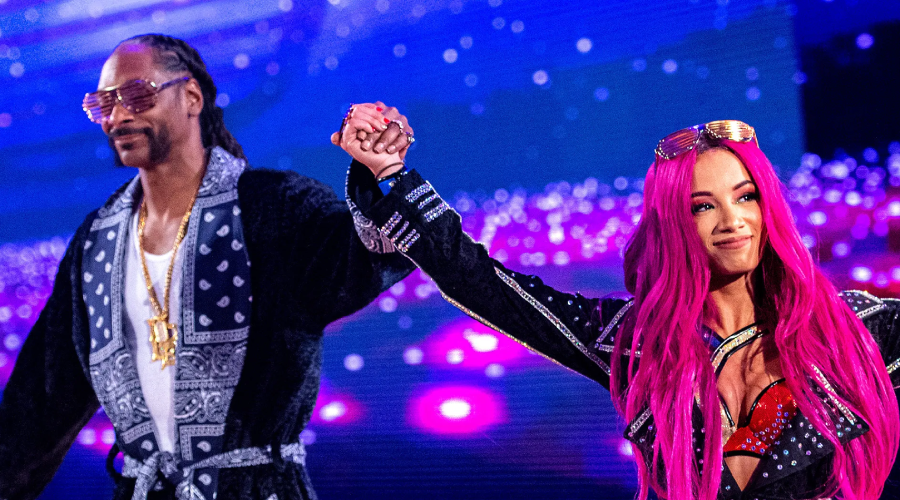 A man in sunglasses and a black patterned robe holds hands with a woman with long bright pink hair, also wearing sunglasses and a decorated outfit, as they raise their arms on a vibrant, purple-lit stage.