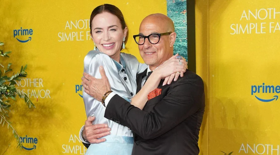 A woman in a light blue dress and a man in a suit and glasses smile and hug on a yellow carpet at an event with "Another Simple Favor" and "prime" logos in the background.