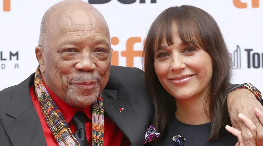 An older man in a red shirt and patterned scarf stands with his arm around a younger woman with straight brown hair and bangs. Both are smiling at an event with a "tiff" logo in the background.