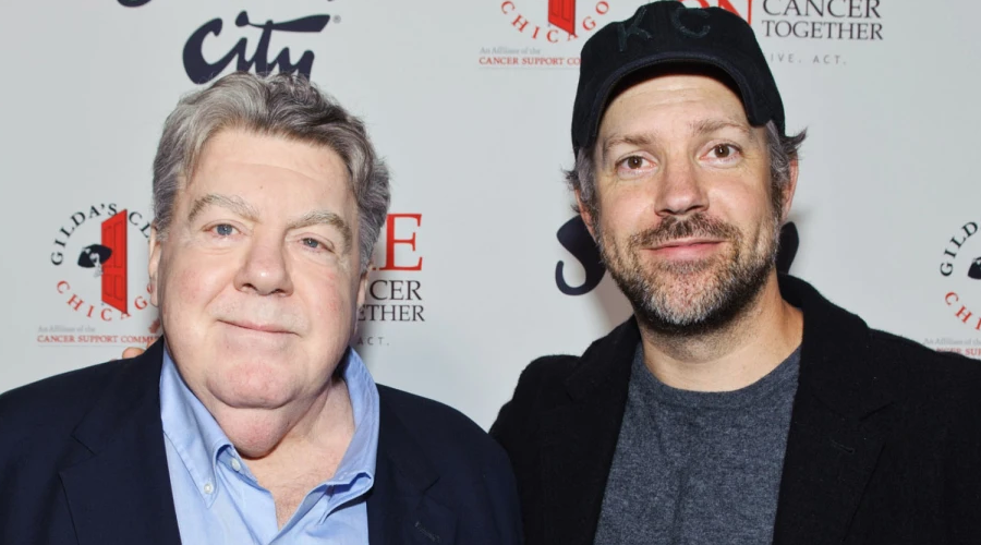 Two men stand side by side at an event with a white step-and-repeat backdrop covered in logos and text. One man has gray hair and wears a suit; the other has a beard and wears a cap and jacket.