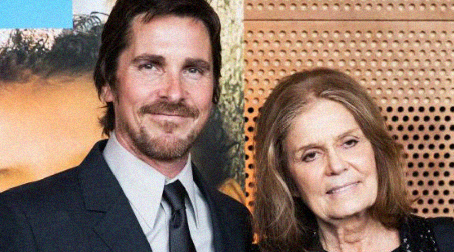 A man in a suit and tie stands next to an older woman with shoulder-length hair, both smiling at the camera against a background with a mix of artwork and wood paneling.