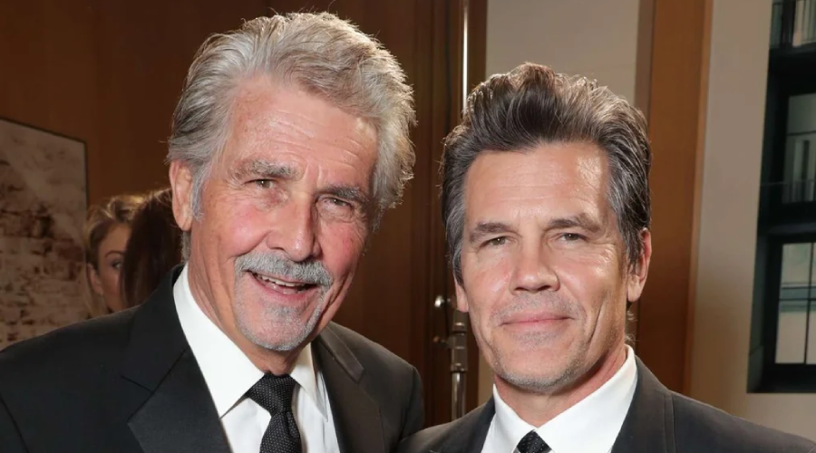 Two men dressed in black suits and ties stand close together, smiling at the camera in a warmly lit indoor setting. Both have gray-streaked hair and are posing for a formal photograph.
