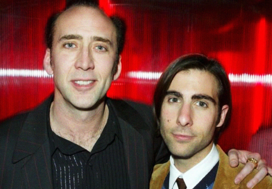 Two men standing close together, one with his arm around the other, both smiling slightly. They are dressed in suits and posed against a red, softly lit background.