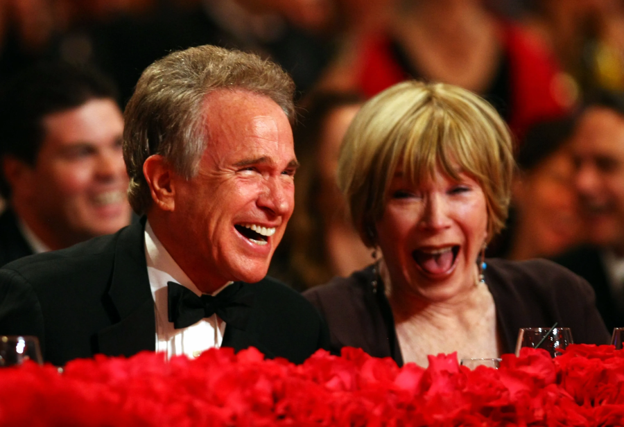 Two people dressed formally sit at a table adorned with red flowers, laughing joyfully at an event. The background shows other smiling guests in a festive atmosphere.