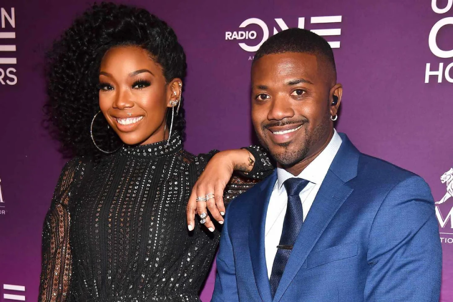 A woman in a black sequined dress with hoop earrings stands beside a man in a blue suit and tie. They are smiling in front of a purple backdrop with white logos and text.
