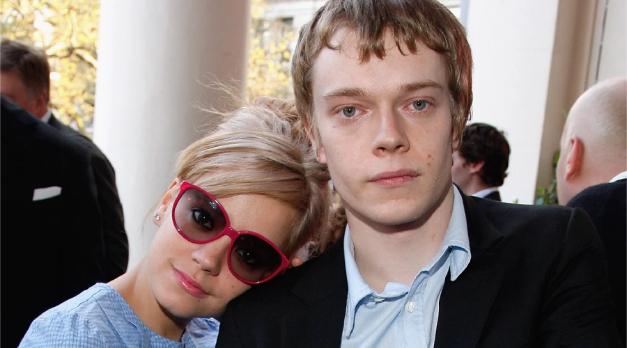 A woman with blond hair and red sunglasses leans her head on the shoulder of a man in a black blazer and blue shirt. They are standing close together outdoors, with other people in the background.