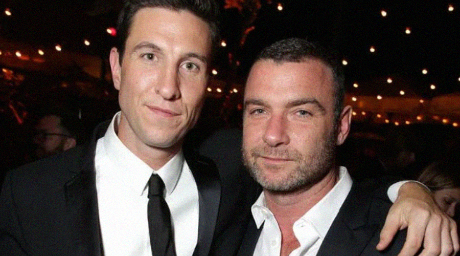 Two men in suits pose closely together and smile at night, under string lights at an outdoor event or party.