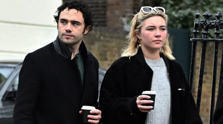 Two people walk outside holding coffee cups. The man has short curly dark hair and wears a black coat. The woman has shoulder-length blonde hair, wears a black jacket, and has sunglasses on her head. Both look ahead.